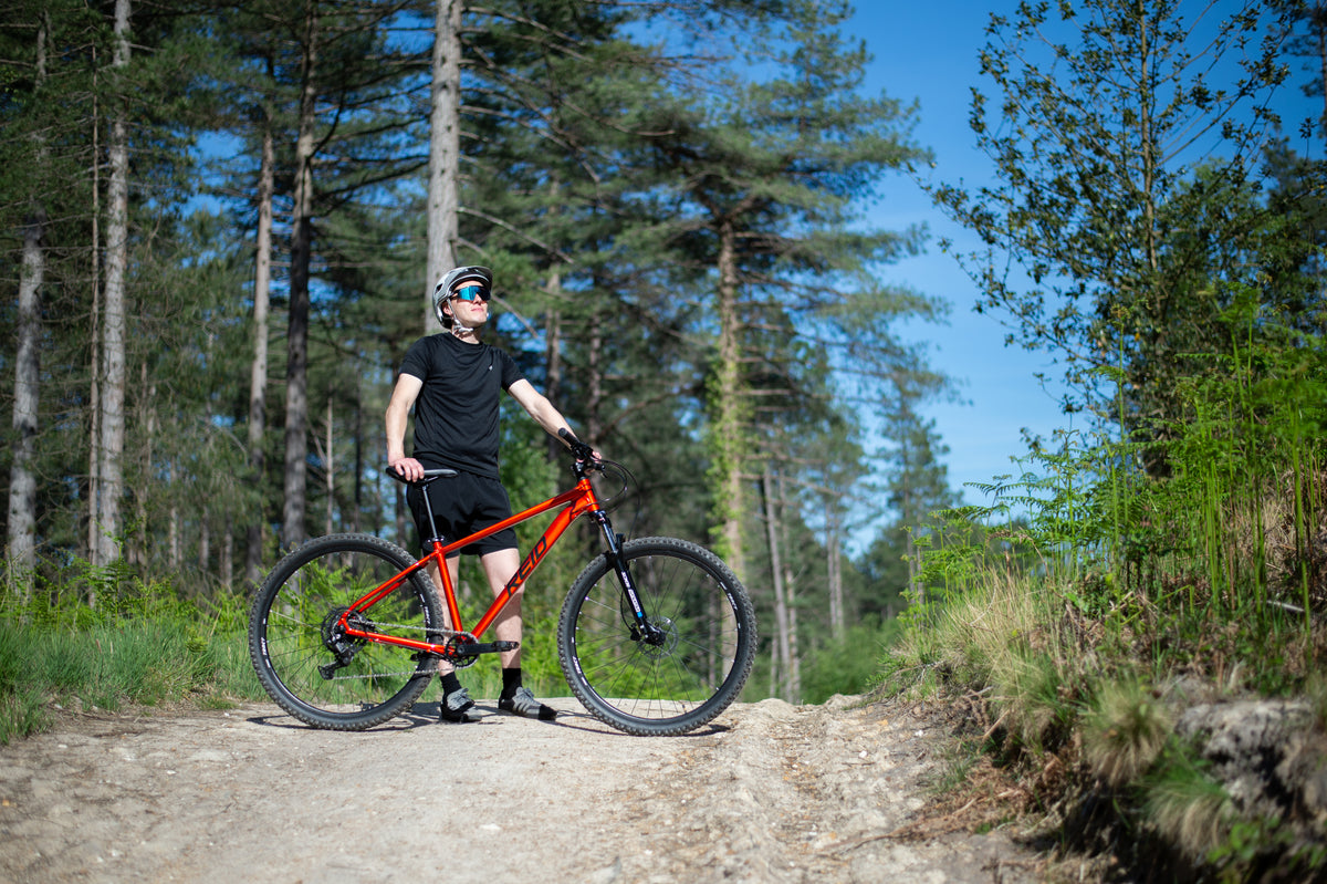 A mountain biker stands on a dirt trail in a forest, holding a red Reid mountain bike. The rider is wearing a black t-shirt, black shorts, and a helmet with blue goggles.