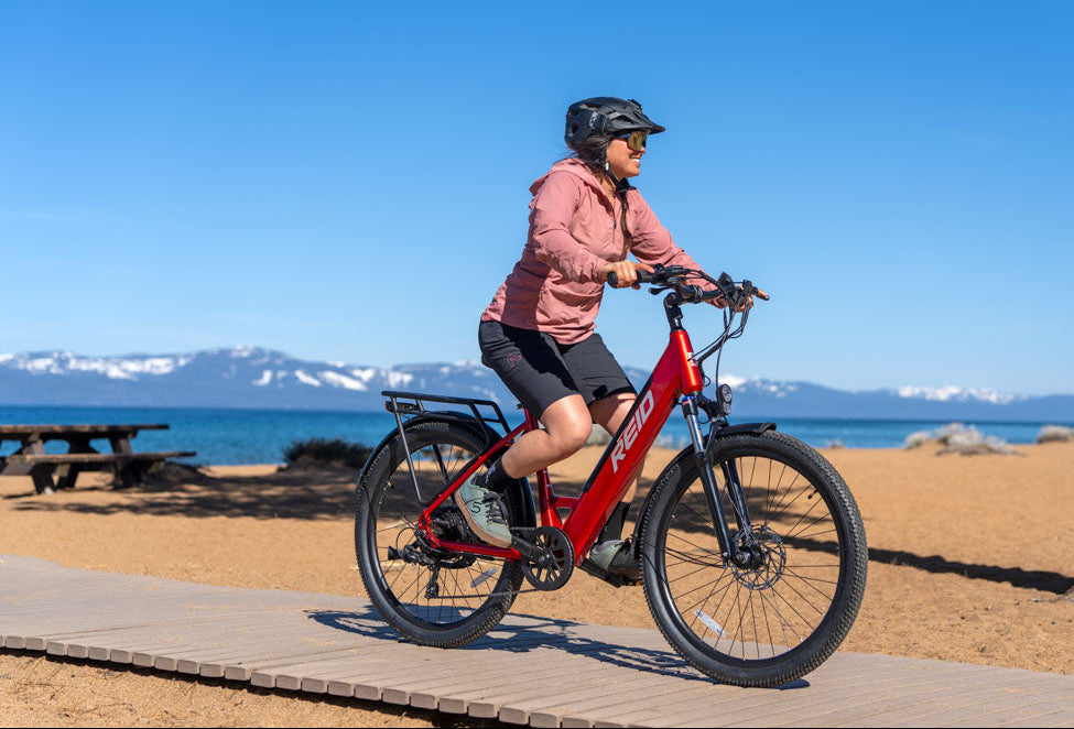 Person riding a red electric Quest 1 ST Reid bike on a scenic beach path with mountains in the background